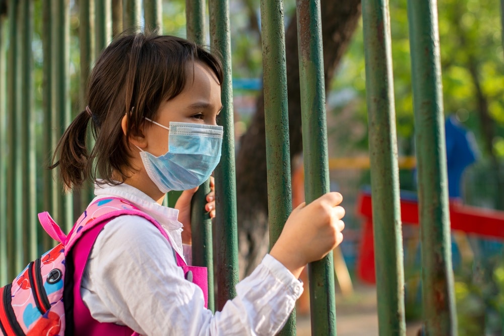 Little school girl standing next to school fence waiting for going back to clases after pandemic outbreak covid